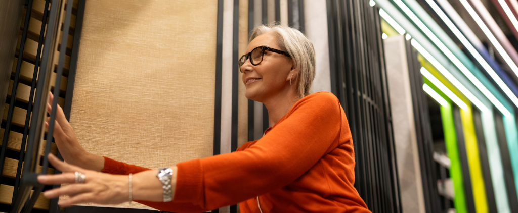 Woman wearing glasses and an orange sweater examines flooring samples displayed on vertical racks inside a modern flooring showroom, with rows of sample boards extending into the background.